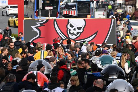 Zahlreiche Menschen laufen an einer Plakatwand mit dem Emblem der Tampa Bay Buccaneers auf dem Odeonsplatz vorbei. 