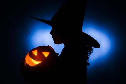 Ein Mädchen im Hexenkostüm hält einen beleuchteten Halloween-Kürbis im Arm.
